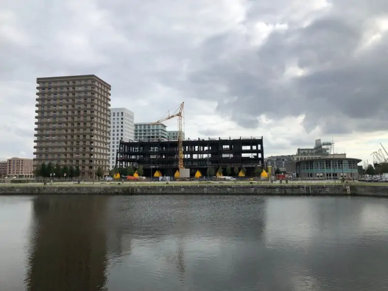 Wide view of the Water Weights load testing project across the canal in Belgium, showing the building under construction alongside the surrounding city skyline
